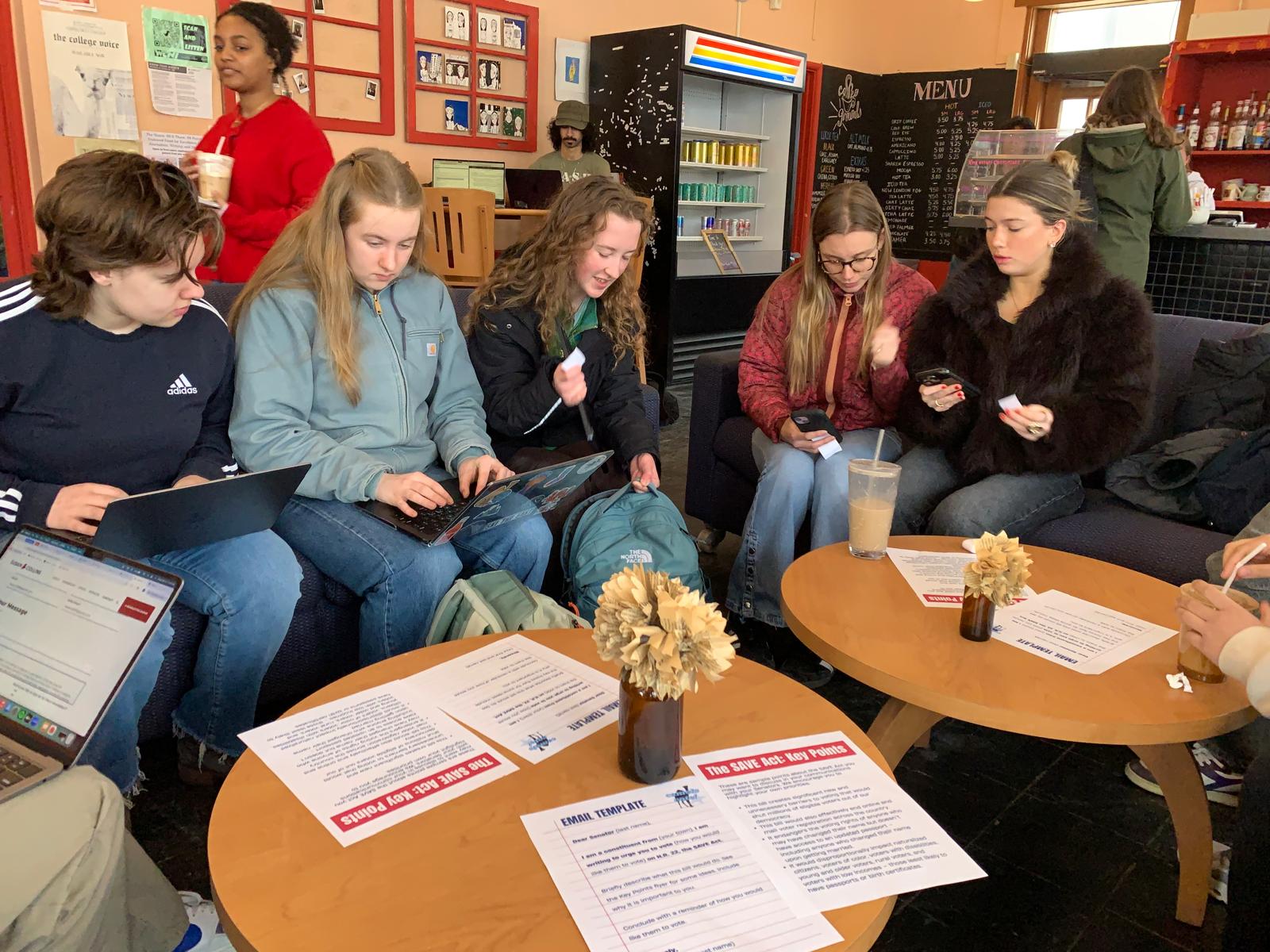 A group of students sitting in a cafe with laptops and SAVE Act key-points handouts spread across the table.